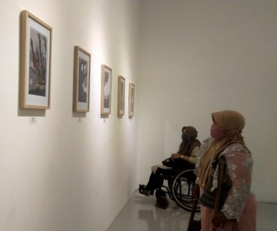 Foto pengguna kursi roda dan pengguna tongkat sedang melihat karya lukis yang tergantung didinding